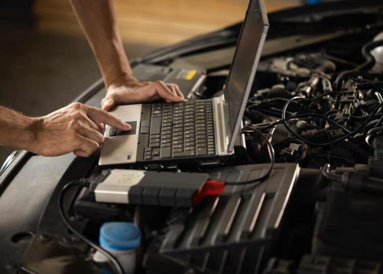 A person uses a laptop atop a car engine bay to run diagnostics with cables and tools connected to the vehicle.