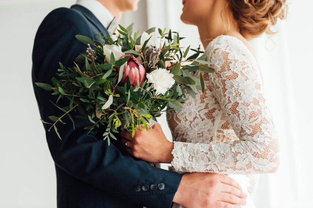 A bride and groom stand together holding a colorful wedding bouquet, posing closely in formal attire.