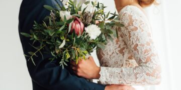 A bride and groom stand together holding a colorful wedding bouquet, posing closely in formal attire.