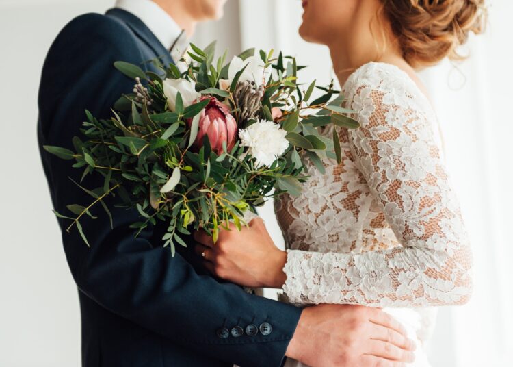 A bride and groom stand together holding a colorful wedding bouquet, posing closely in formal attire.