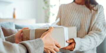 A smiling woman is holding a wrapped present in her hands as someone gives it to her.