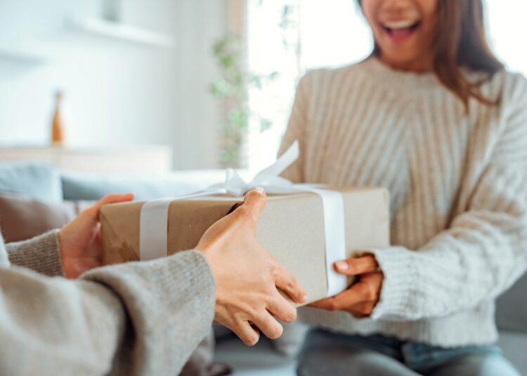 A smiling woman is holding a wrapped present in her hands as someone gives it to her.