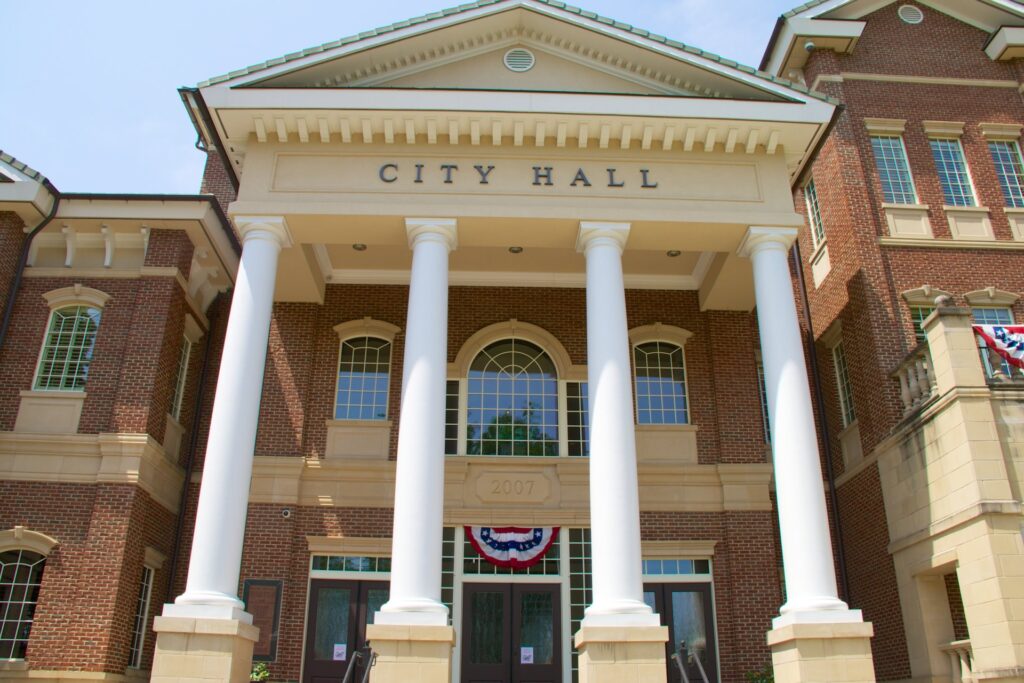 The entrance of a city hall building. It features four massive white columns and red brick siding.