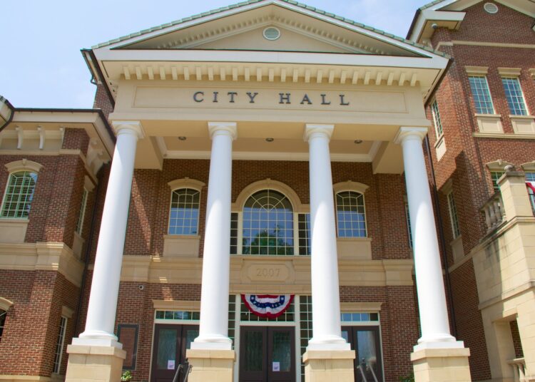 The entrance of a city hall building. It features four massive white columns and red brick siding.