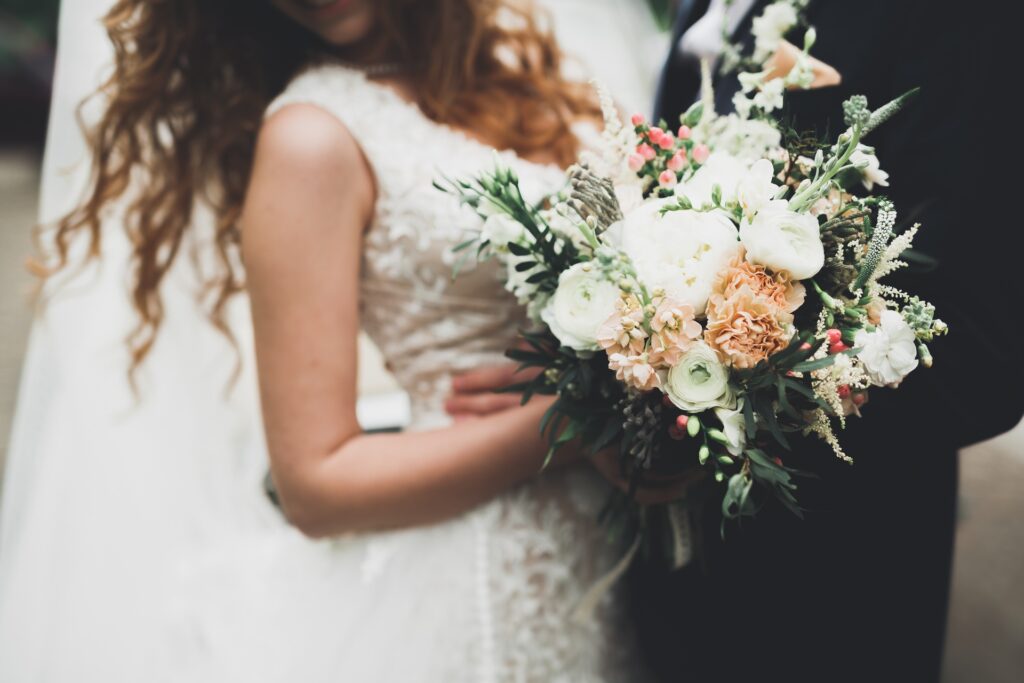 A woman in a white lace gown carries a mixed bouquet of light flowers and greenery during a wedding.