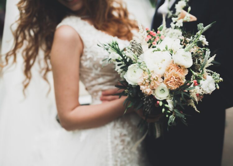 A woman in a white lace gown carries a mixed bouquet of light flowers and greenery during a wedding.