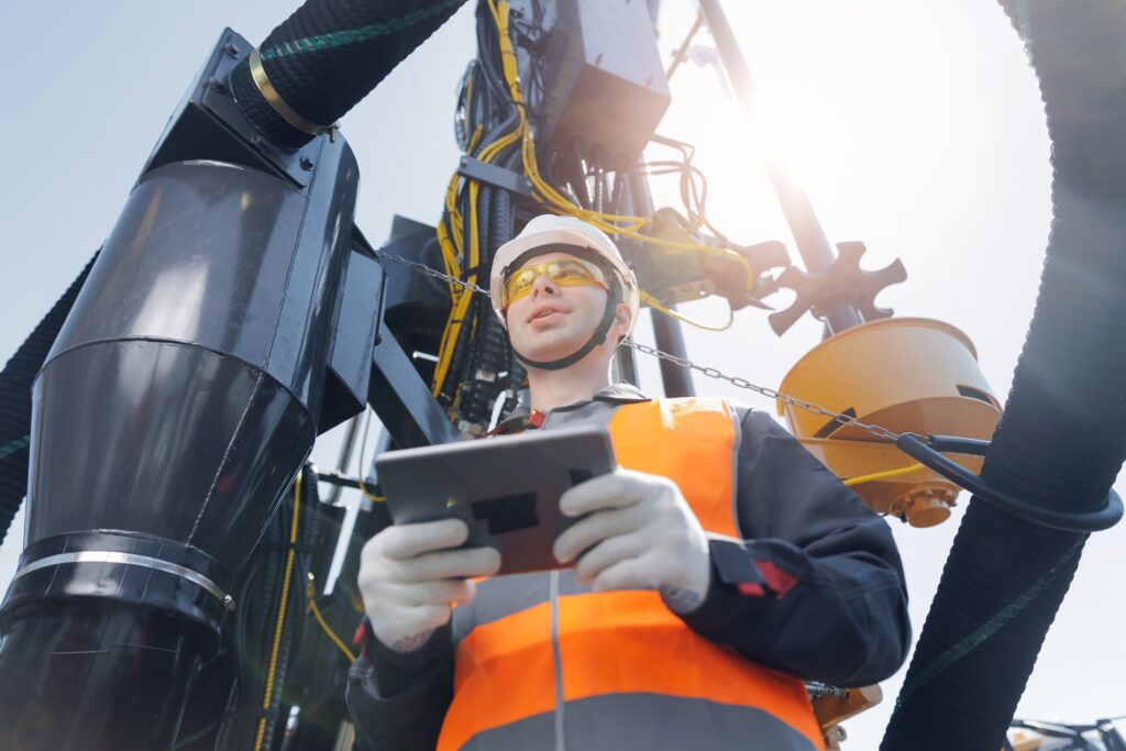 An industrial operator wearing PPE holds a tablet to control the drilling rig system sitting behind him.