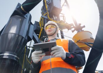 An industrial operator wearing PPE holds a tablet to control the drilling rig system sitting behind him.