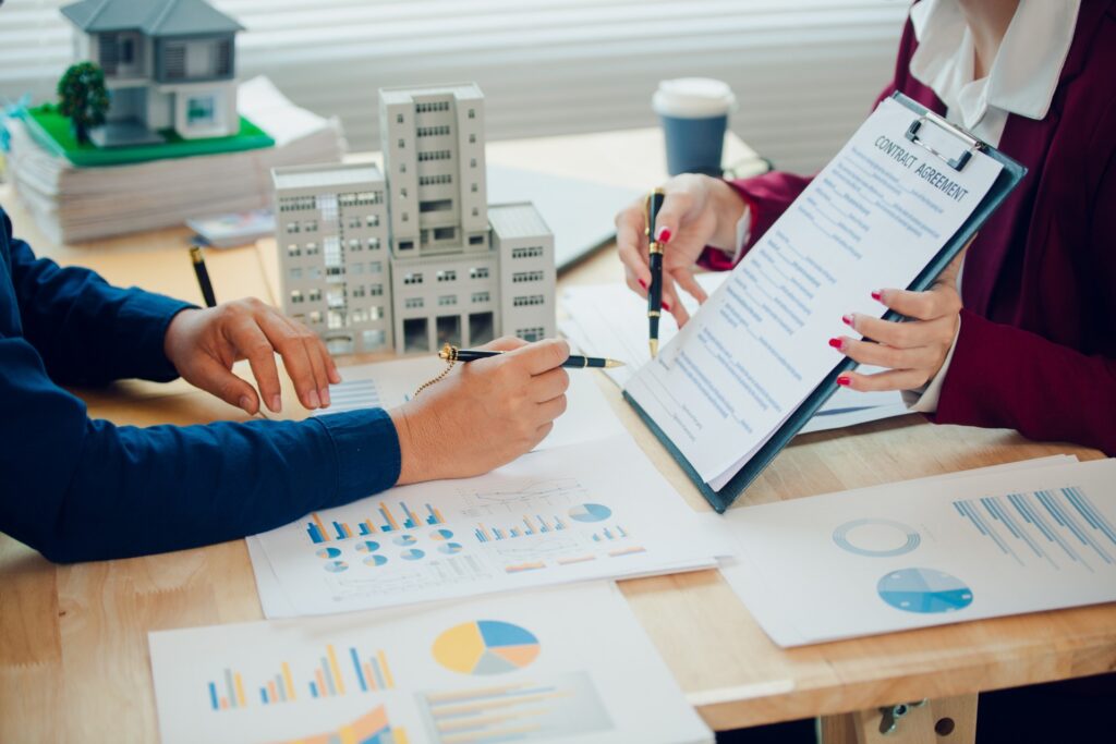 Two people are sitting across from each other at a desk. The desk is cluttered with papers.