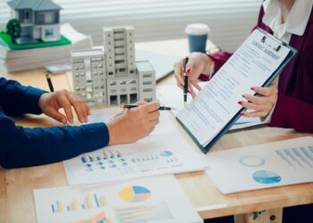 Two people are sitting across from each other at a desk. The desk is cluttered with papers.