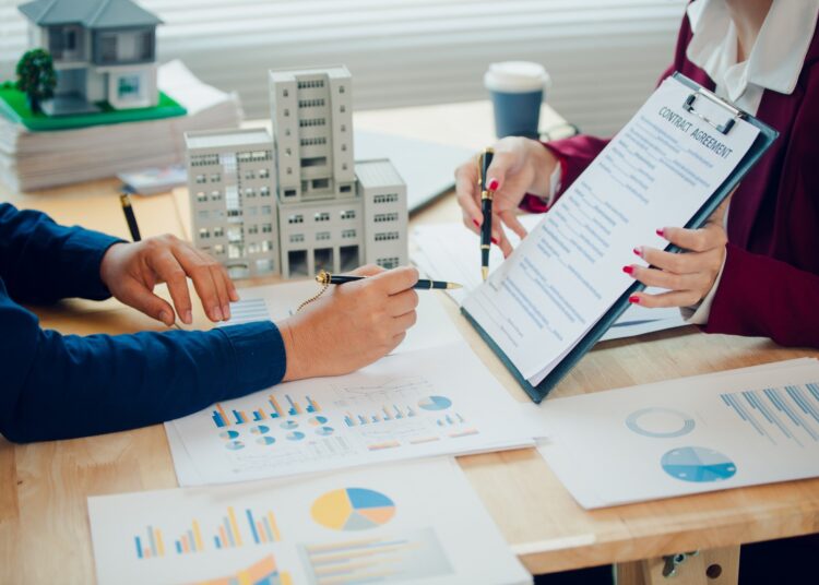 Two people are sitting across from each other at a desk. The desk is cluttered with papers.