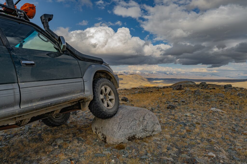 An off-roading car parked on a rocky trail with big clouds overhead. The car’s right front tire is parked on a big rock.