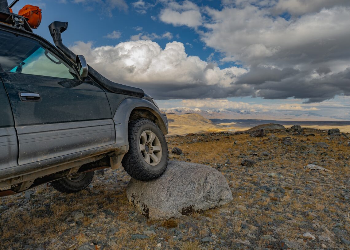 An off-roading car parked on a rocky trail with big clouds overhead. The car’s right front tire is parked on a big rock.