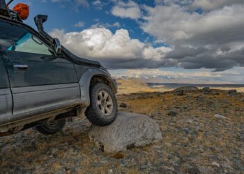An off-roading car parked on a rocky trail with big clouds overhead. The car’s right front tire is parked on a big rock.