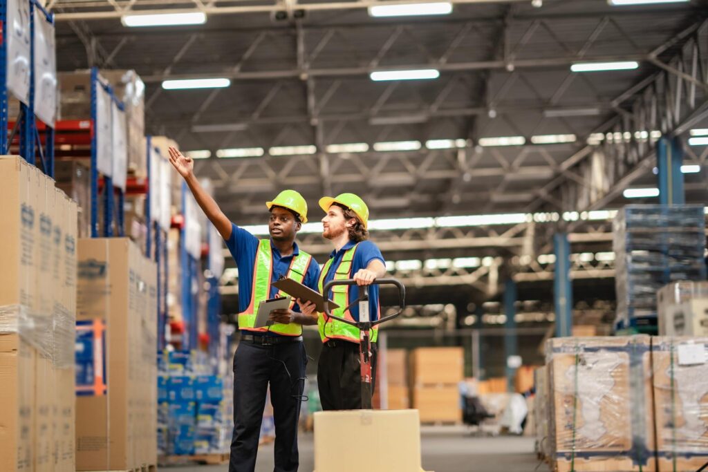 Two warehouse workers are discussing the facility layout, tablets in hand. They both have hard hats and safety vests on.