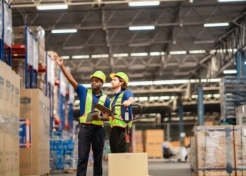 Two warehouse workers are discussing the facility layout, tablets in hand. They both have hard hats and safety vests on.