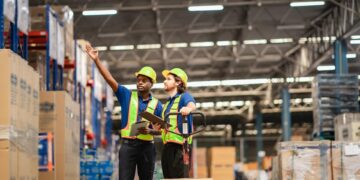 Two warehouse workers are discussing the facility layout, tablets in hand. They both have hard hats and safety vests on.