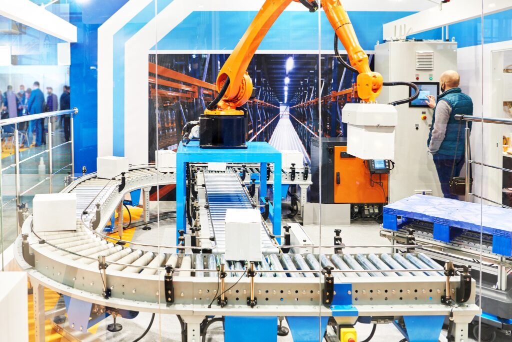 A robotic palletizing arm loads boxes on a conveyor system while an operator controls machinery in a modern warehouse setup.