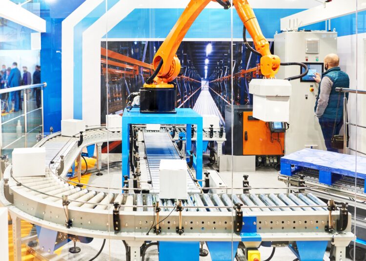 A robotic palletizing arm loads boxes on a conveyor system while an operator controls machinery in a modern warehouse setup.