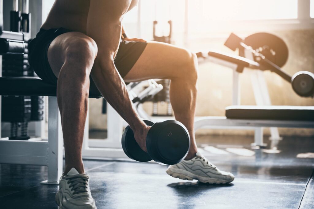 A person holds a dumbbell near the floor while leaning forward. There are gym equipment and weights in the background.