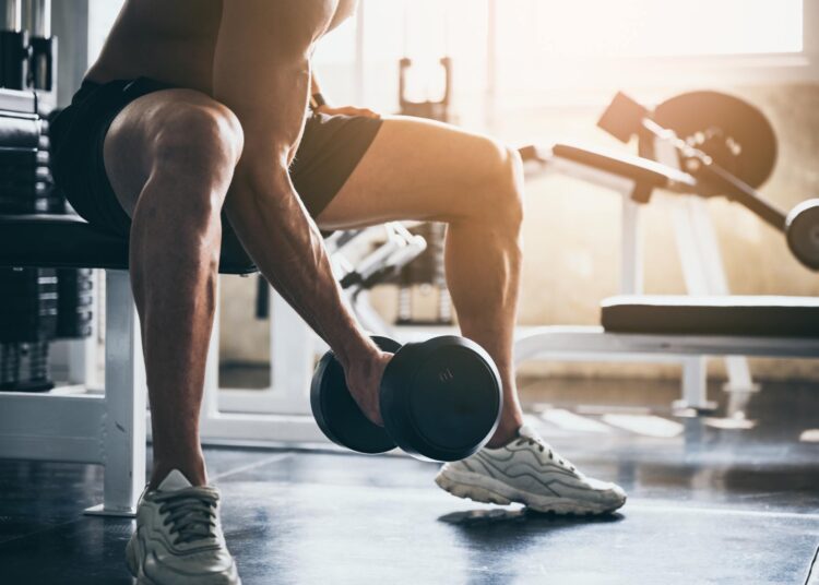 A person holds a dumbbell near the floor while leaning forward. There are gym equipment and weights in the background.