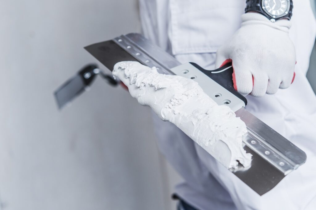 A person in white protective gear holding a drywall knife with mud in one hand and a smaller clean knife in the other.