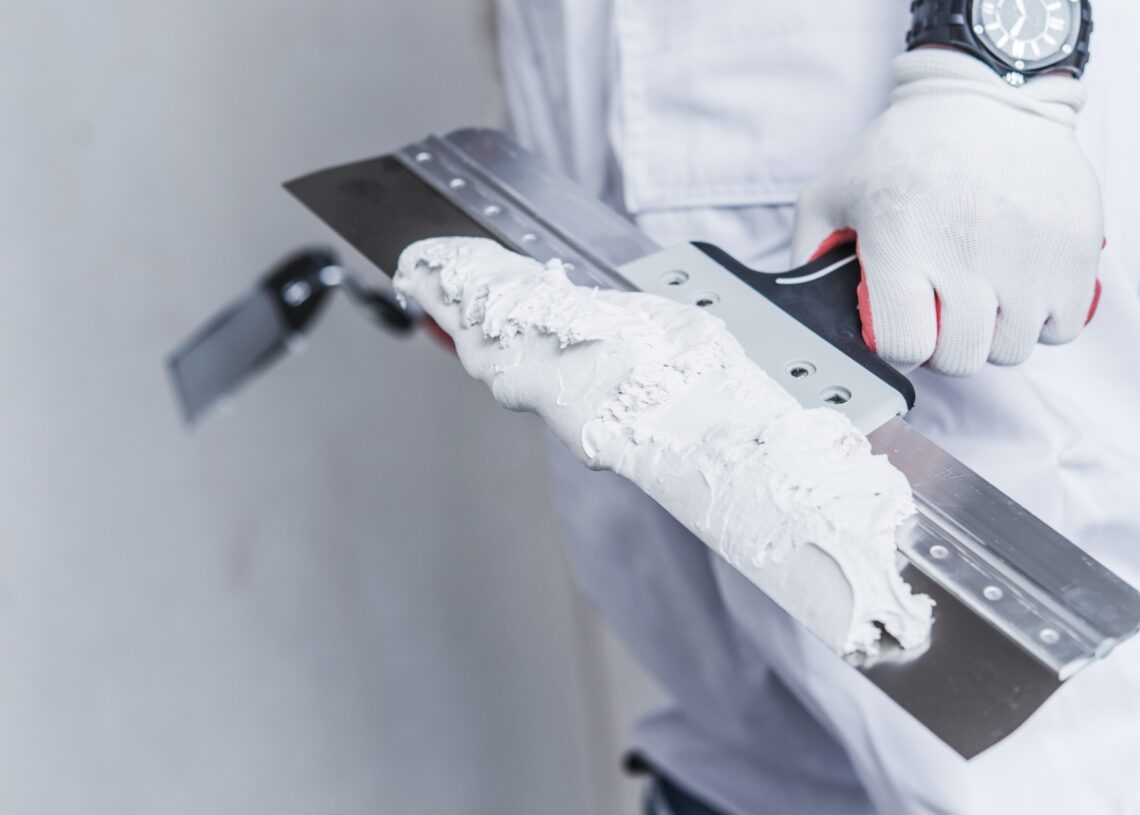 A person in white protective gear holding a drywall knife with mud in one hand and a smaller clean knife in the other.