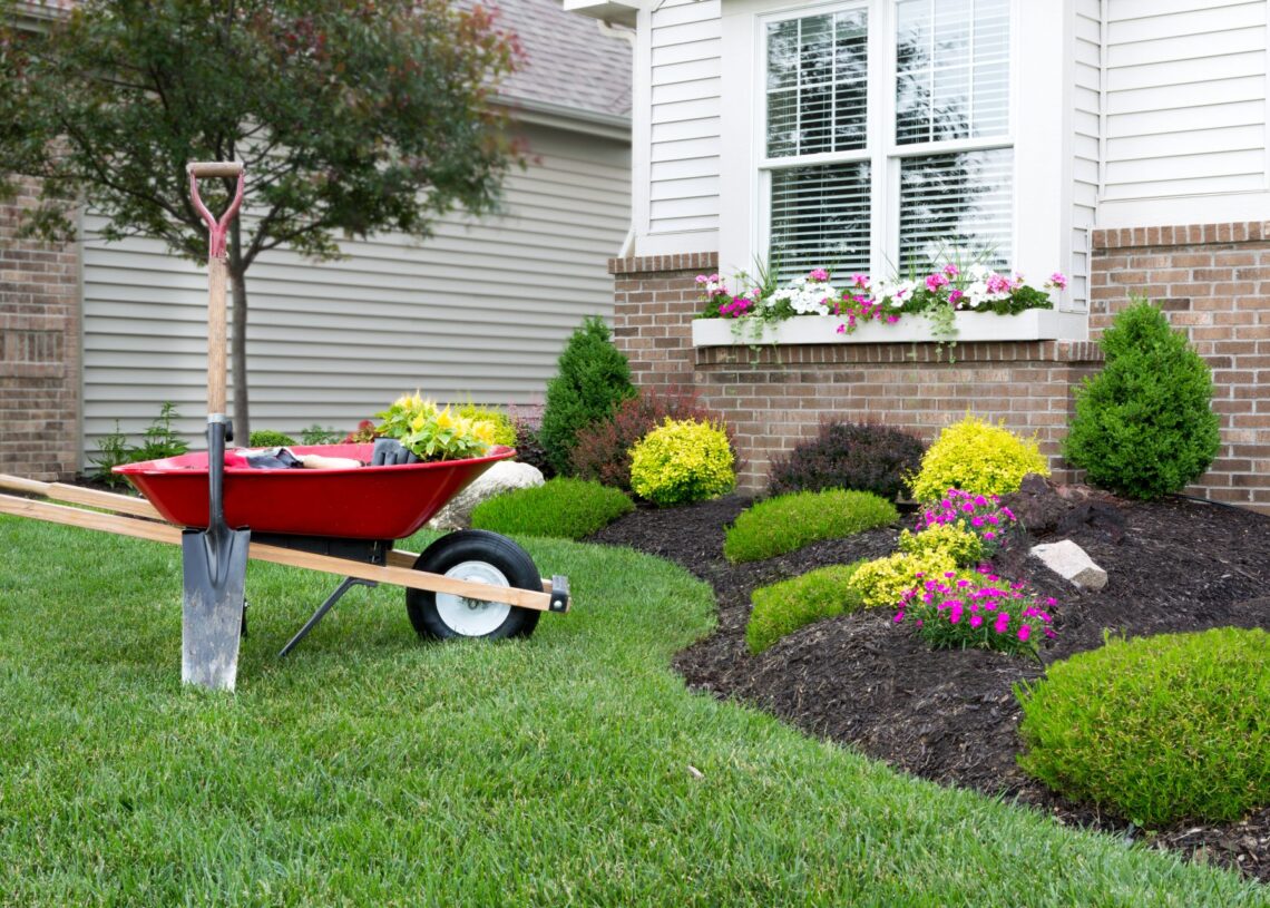 A red wheel-barrow and a shovel sit next to freshly landscaped garden boxes in the front of a house.