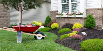 A red wheel-barrow and a shovel sit next to freshly landscaped garden boxes in the front of a house.