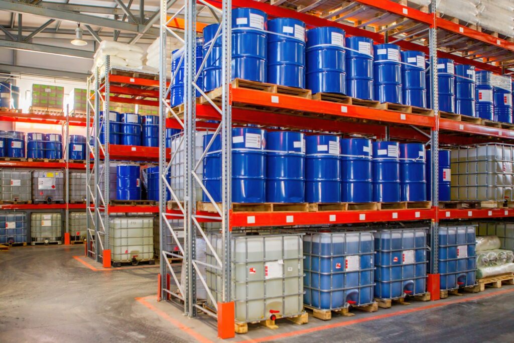 Industrial storage shelves contain blue steel drums on the top shelves and plastic square containers on the bottom.