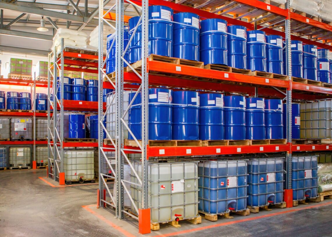 Industrial storage shelves contain blue steel drums on the top shelves and plastic square containers on the bottom.