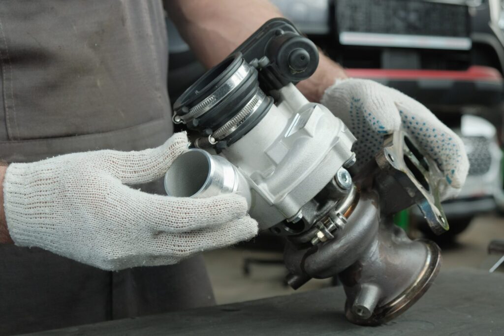 A person holding a metal turbocharger part in a workshop, with a vehicle engine and tools visible nearby.