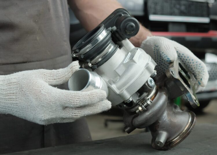 A person holding a metal turbocharger part in a workshop, with a vehicle engine and tools visible nearby.