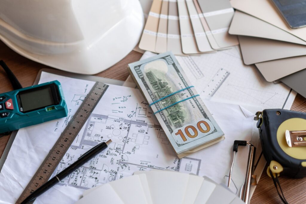 A wooden table with various construction plans and supplies, like a ruler and a hard hat, along with a stack of $100 bills.