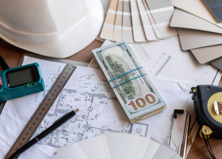A wooden table with various construction plans and supplies, like a ruler and a hard hat, along with a stack of $100 bills.