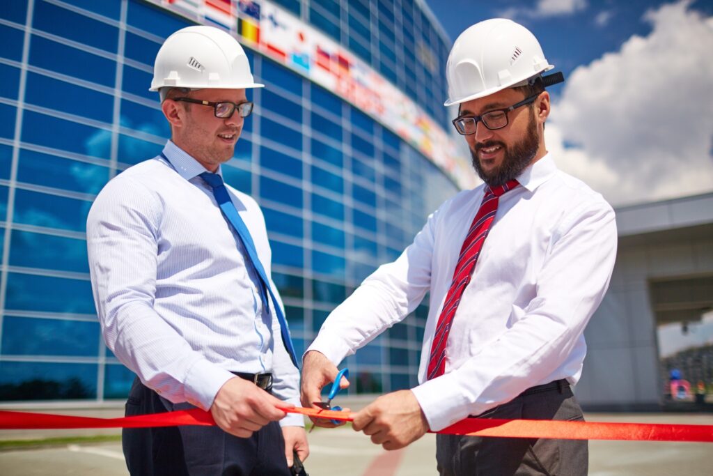 Two men stand outside a new building and perform a ribbon-cutting ceremony. Both wear hard hats and ties.