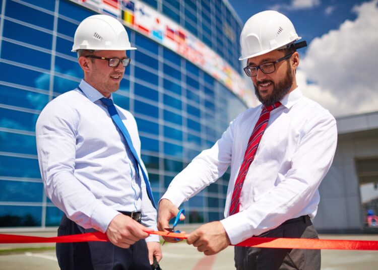 Two men stand outside a new building and perform a ribbon-cutting ceremony. Both wear hard hats and ties.