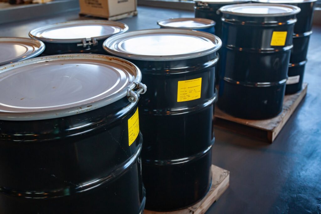 Large, black metal drums sit in groups of four on wooden pallets in a warehouse. Each drum features a yellow sticker.