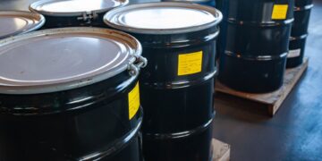 Large, black metal drums sit in groups of four on wooden pallets in a warehouse. Each drum features a yellow sticker.