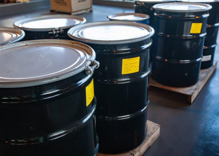 Large, black metal drums sit in groups of four on wooden pallets in a warehouse. Each drum features a yellow sticker.