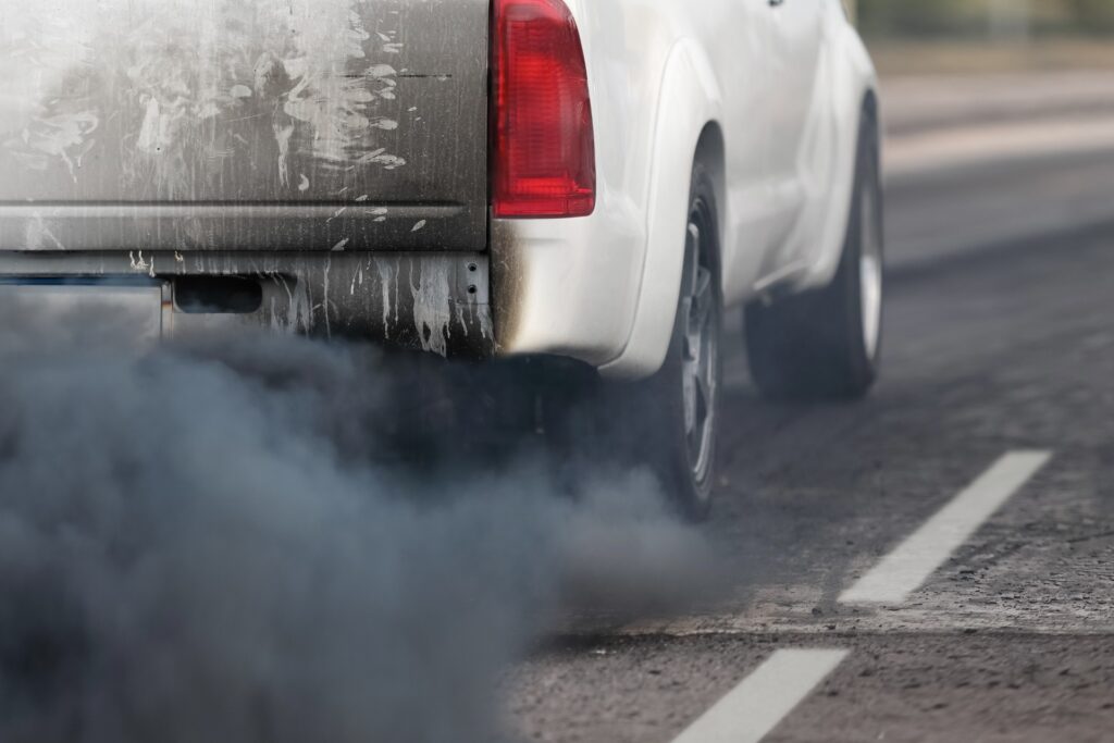 A white pickup truck driving on a road while emitting thick black exhaust smoke from the tailpipe.