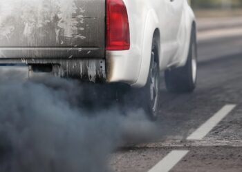 A white pickup truck driving on a road while emitting thick black exhaust smoke from the tailpipe.