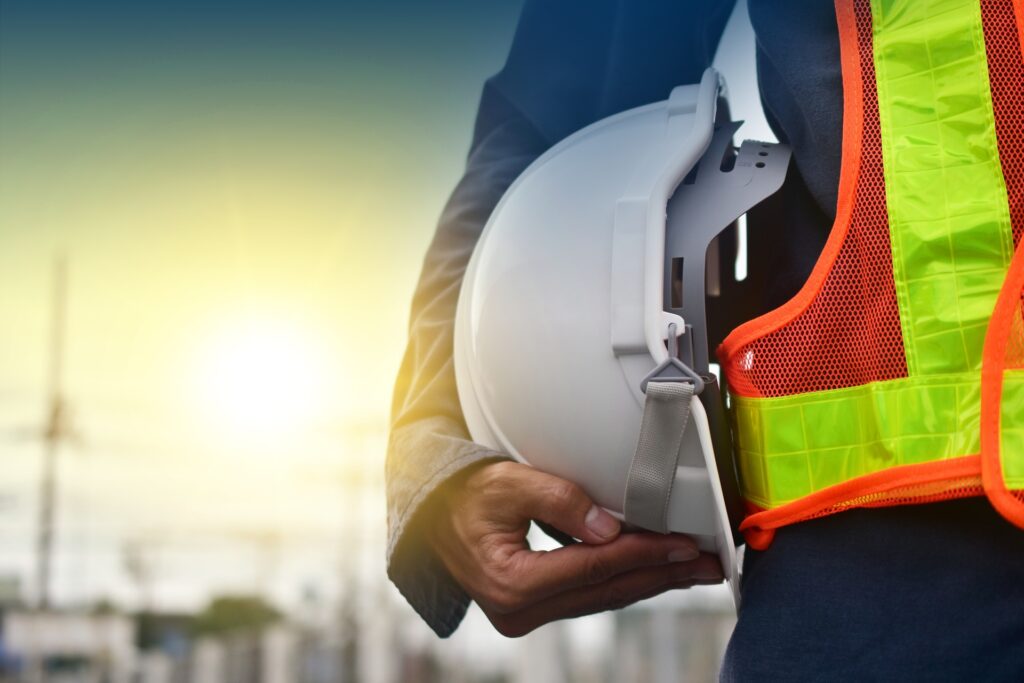 The body of a construction worker wearing an orange safety vest and holding a hard hat set against the setting sun.