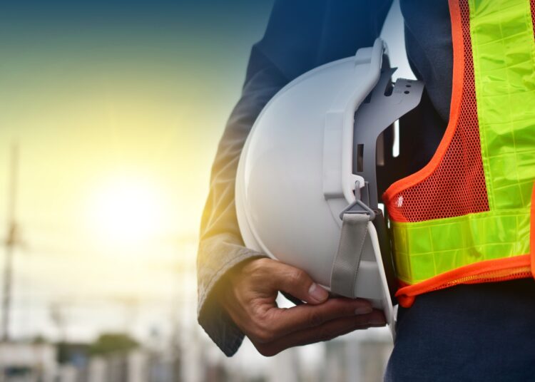 The body of a construction worker wearing an orange safety vest and holding a hard hat set against the setting sun.