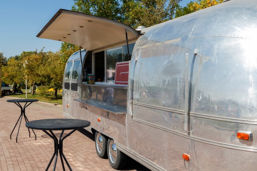 A close-up of a silver trailer parked next to two outdoor tables. The side of the trailer is open, showing an ordering window.