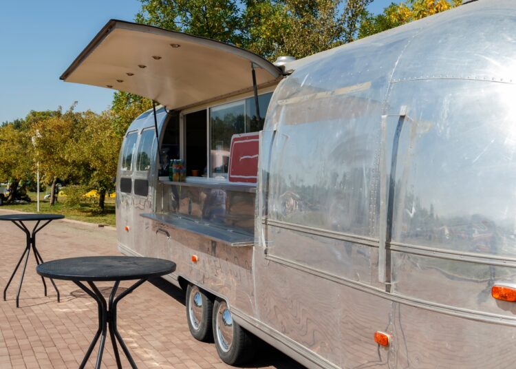 A close-up of a silver trailer parked next to two outdoor tables. The side of the trailer is open, showing an ordering window.