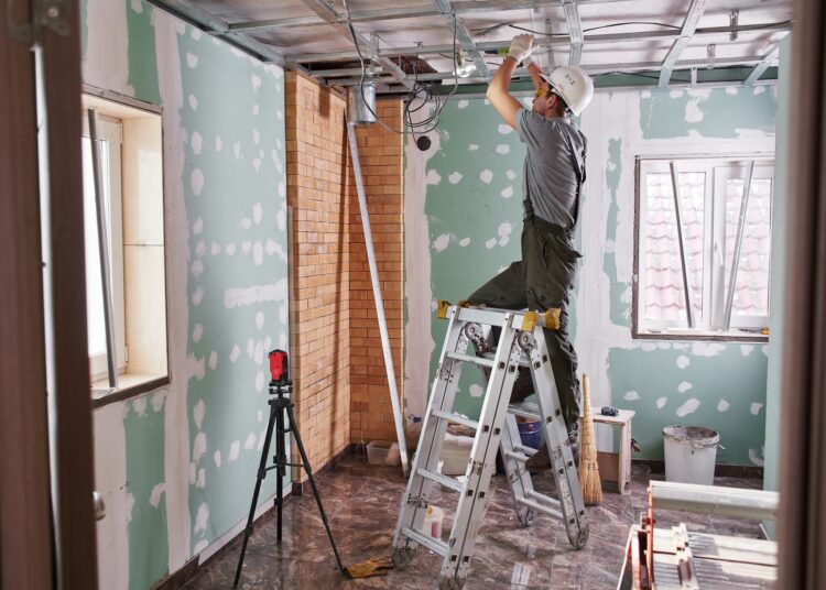A man in overalls and a hard hat is standing on a step ladder, working on some wiring that is on the ceiling of the room.
