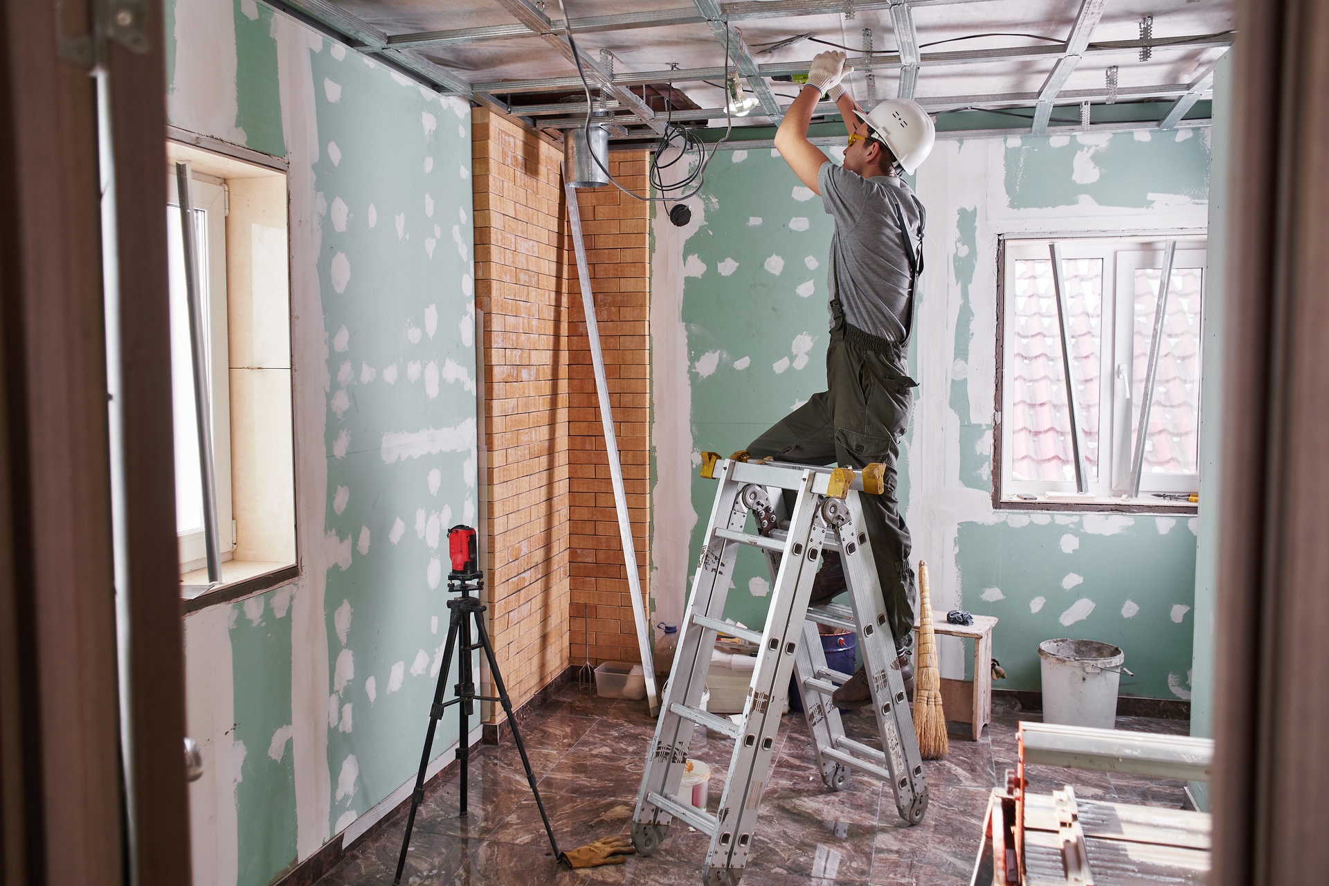 A man in overalls and a hard hat is standing on a step ladder, working on some wiring that is on the ceiling of the room.