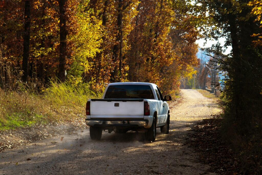 A white pickup truck is driving through the woods on a dirt road. The leaves on the trees are changing colors.