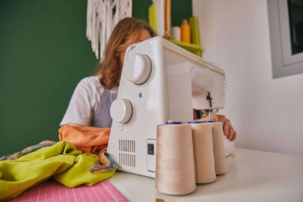 A person sewing at a white sewing machine with thread spools, bright fabric, and cutting mat on a tidy craft workspace.